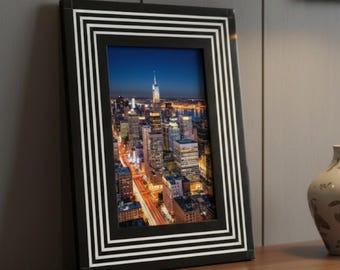 Puede incluir: Un marco de fotos negro con un borde a rayas blancas, que muestra un paisaje urbano nocturno con edificios iluminados y rastros de luces de vehículos. El marco está sobre una superficie de madera.