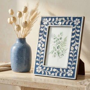 Peut inclure: Cadre photo décoratif bleu avec incrustations florales blanches. Il contient une impression botanique de feuilles vertes. Un vase bleu avec des fleurs séchées est à gauche. Un élément de décoration pour la maison.