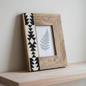 Op de afbeelding: Een houten fotolijst met een zwart-wit geometrisch ontwerp aan de linkerkant. De lijst bevat een zwart-witte tekening van een varenblad. Hij staat op een lichtgekleurde houten plank.