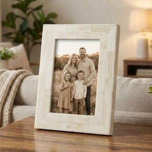 Puede incluir: Un marco de fotos rectangular blanquecino con un retrato familiar en su interior. El marco tiene un diseño texturizado, similar a un mosaico. La foto muestra a una familia de cuatro personas de pie en un campo.