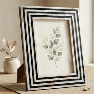 Peut inclure: Un cadre photo rectangulaire doté d'une bordure géométrique composée de bandes noires alternées et de carreaux de nacre blanche irisée. Le cadre contient une illustration botanique de tiges feuillues aux tons verts et beiges.