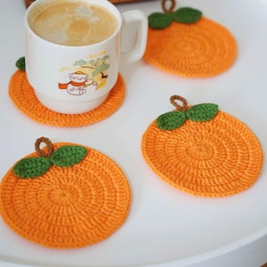Puede incluir: Una taza blanca con un diseño de dibujos animados se encuentra sobre un posavasos naranja de ganchillo con forma de naranja. También se ven tres posavasos naranjas adicionales con hojas verdes y tallos marrones.
