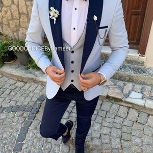 May include: A light blue suit with navy blue lapels and a matching bow tie. The suit includes a waistcoat, dress trousers, and black dress shoes. A small floral buttonhole and a chain with a decorative pin are attached to the jacket.