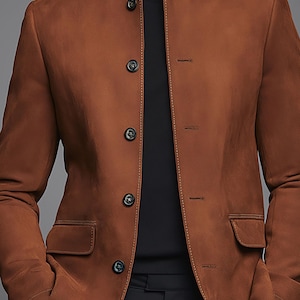 Może przedstawiać: Brązowa zamszowa kurtka z kołnierzem mandaryńskim i czterema guzikami. Kurtka ma dwie przednie kieszenie i jest noszona na czarnym golfie. Kurtka ma ciepły, neutralny kolor.