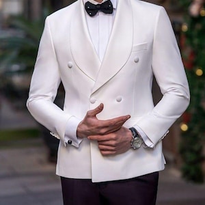 Może przedstawiać: Biała dwurzędowa marynarka z czarną muchą. Marynarka ma klapy z wycięciami i dwa rzędy guzików. Tekst "GOODCHOICEBYSameer" znajduje się na górze. Widoczny jest zegarek z czarnym skórzanym paskiem.