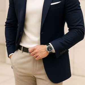 Może przedstawiać: Granatowa marynarka z białą poszetką, w połączeniu z kremowym golfem i beżowymi spodniami. Czarny pasek i zegarek na rękę z czarnym skórzanym paskiem dopełniają stylizację. Widoczny jest napis "GOODCHOICEBYSameer".