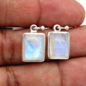Puede incluir: Un par de pendientes rectangulares con piedras de luna facetadas engastadas en marcos de plata. Cada piedra muestra una apariencia blanca translúcida con un brillo iridiscente azul distintivo. Los pendientes están sujetos a ganchos de plata.