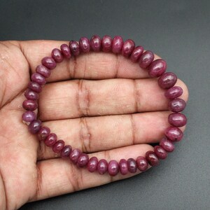Peut inclure: Un bracelet de pierres précieuses rubis. Le bracelet est composé de nombreux petits rubis ovales de couleur rouge foncé. Les pierres précieuses sont enfilées pour former un cercle complet. Le bracelet est présenté sur un fond sombre. Les pierres ont une surface lisse et polie.