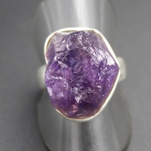 Op de afbeelding: Een zilveren ring met een grote, ruwe amethist edelsteen. De amethist heeft een dieppaarse kleur met een ruw, getextureerd oppervlak. De ring heeft een eenvoudig ontwerp, met de edelsteen gezet in een zilveren bezel.