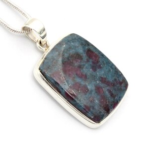 May include: A rectangular pendant featuring a polished, mottled stone with deep teal and dark magenta patches, set in a smooth silver bezel. The pendant hangs from a silver snake chain, showcasing a clean, metallic finish and a polished stone surface.