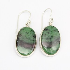 Könnte beinhalten: Ein Paar oval geformte Ohrringe mit polierten grünen und schwarzen Edelstein-Cabochons, die in silberfarbene Metallfassungen eingefasst sind. Jeder Ohrring hängt an einem einfachen silberfarbenen Haken und zeigt die natürliche Maserung.