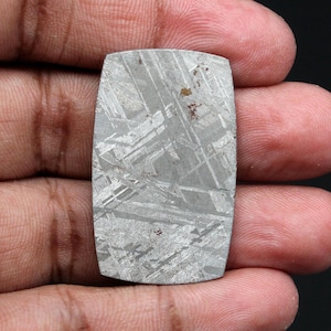 Op de afbeelding: Een rechthoekige, gepolijste, grijze edelsteen met een uniek kruispatroon. De steen heeft afgeronde hoeken en een licht getextureerd oppervlak. De edelsteen wordt in een hand gehouden.