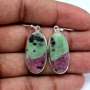 Puede incluir: Un par de pendientes ovalados con un marco de metal plateado. Los pendientes presentan una piedra natural con una parte superior verde, una base rosa y motas negras. Los pendientes se sostienen en una mano.
