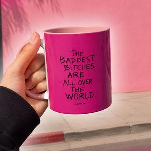 Peut inclure: Un mug en céramique rose vif avec un intérieur et une anse rose clair. L'extérieur présente un texte noir qui dit, The BADDEST BITCHES ARE ALL OVER THE WORLD, avec le mot ADELA imprimé en plus petites lettres noires sous le texte principal.