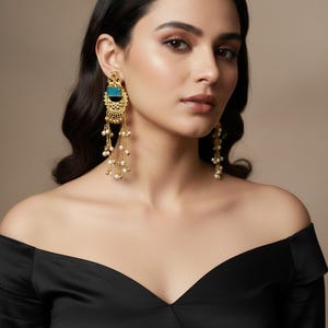 Könnte beinhalten: Goldfarbene Ohrringe mit einem türkisfarbenen quadratischen Mittelteil und baumelnden Perlenakzenten. Die Ohrringe haben ein verziertes Filigran-Design und werden von einer Person getragen, die ein schwarzes schulterfreies Oberteil trägt.