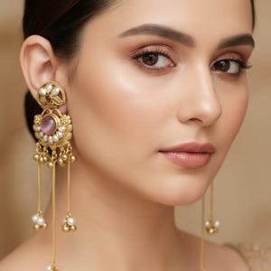 Puede incluir: Primer plano de una mujer con pendientes dorados ornamentados. Los pendientes presentan un diseño floral con una piedra central púrpura, rodeada de perlas y cadenas doradas colgantes con detalles de perlas y cuentas doradas. La mujer tiene el pelo oscuro y mira a la cámara.