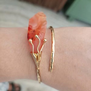 Può includere: Un bracciale aperto color oro con una pietra lucida traslucida color rosso-arancio, incastonata in una montatura metallica che ricorda un bocciolo di fiore. Il bracciale presenta una finitura martellata e testurizzata con design aperto.