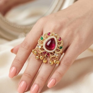 Peut inclure: Une bague imposante dorée ornée d'une grande pierre centrale rouge en forme de goutte, entourée de petites perles blanches. La monture en métal travaillé comprend des pierres rouges et vertes et des breloques sphériques dorées.