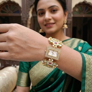 May include: A gold-tone rectangular wristwatch featuring a white dial with black numerals. The watch band is composed of ornate, decorative links embellished with clear stones, small white pearls, and central red accents, creating a jewelry-style timepiece.