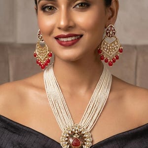 May include: A woman wearing a multi-strand pearl necklace with a large pendant and matching earrings. The pendant and earrings feature gold filigree, red accents, and small red bead drops. The woman is wearing a black top.