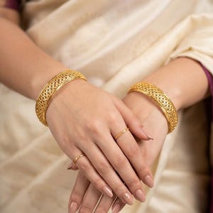May include: Two gold bangles featuring an intricate, openwork lattice pattern with a textured, rope-like border. The bangles have a polished, reflective finish. Additionally, two thin, matching gold rings are worn on the fingers, completing the set.