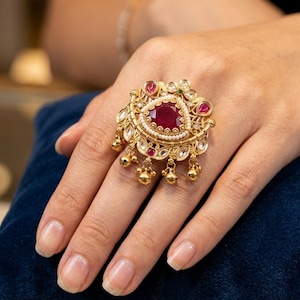 Puede incluir: Un anillo dorado con una gran piedra preciosa roja oscura ovalada en el centro, rodeada de piedras rojas y transparentes más pequeñas. El anillo tiene un diseño detallado y ornamentado con detalles dorados colgantes y pequeñas perlas. El anillo se muestra en una mano.