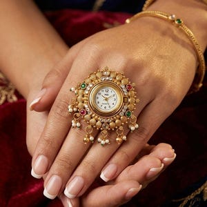 May include: A gold-toned ring watch featuring a round white analog clock face with black numerals. The circular frame is decorated with small clear stones, red and green accents, and dangling white pearl-like beads in a textured metal setting.