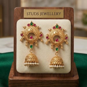 May include: Gold earrings with a sunburst design, featuring red and green gemstones. The earrings have a bell-shaped drop adorned with small pearls. The display stand has a gold sign that reads "STUDS JEWELLERY".