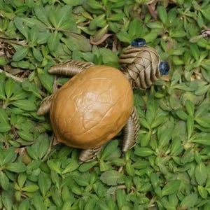 May include: A small turtle figurine crafted with a brown walnut shell as the body and coiled metallic wire forming the head, four legs, and tail. The head features two small, dark blue circular beads serving as eyes, attached to the wire structure.
