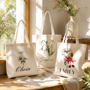 Puede incluir: Tres bolsas de tela de color blanco roto con ilustraciones florales y nombres personalizados. La bolsa de la izquierda muestra margaritas y el nombre Olivia, la central flores de nieve y el nombre Sophia, y la de la derecha flores rosas y el nombre EMILY.