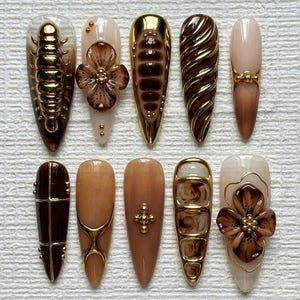 Puede incluir: Una colección de diez uñas artificiales largas en forma de almendra. Las uñas presentan varios diseños, incluyendo detalles dorados, adornos florales y patrones texturizados en tonos marrones, beige y dorados. Algunas uñas tienen un acabado brillante, otras son mate.