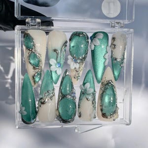 Puede incluir: Un juego de diez uñas artificiales en una caja de plástico transparente. Las uñas presentan una variedad de diseños, que incluyen colores turquesa y blanco, acentos florales y adornos plateados. Las uñas tienen varias formas, incluyendo almendra y estilete.
