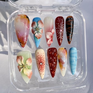 Puede incluir: Un juego de diez uñas artificiales en una caja de plástico transparente. Las uñas presentan varios diseños, incluyendo adornos florales, patrones abstractos y degradados en colores como rosa, azul, rojo y naranja. Algunas uñas tienen un acabado brillante.