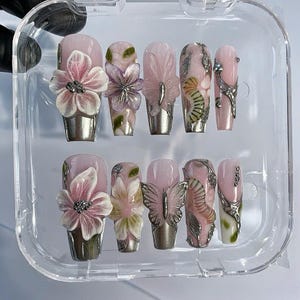 May include: A set of ten artificial nails in a clear plastic case. The nails are light pink with 3D floral, butterfly, and leaf designs, and silver metallic accents. Some nails have rhinestones.