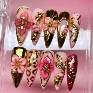 May include: A collection of ten artificial nails with diverse designs. The nails incorporate pink, gold, and clear elements. Some nails feature 3D floral designs, while others display gold embellishments and leopard print patterns. The nails are presented on a pink surface.