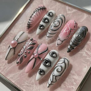 May include: A set of ten long, almond-shaped artificial nails. Designs include pink and white bases with silver embellishments, including eye motifs, swirls, and floral patterns. The nails are arranged on a pink surface within a silver-edged frame.