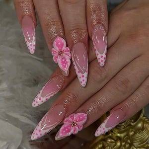 Op de afbeelding: Set van lange, puntige acrylnagels. De nagels hebben een roze en witte kleurstelling met een French tip design. Sommige nagels hebben een wit stippenpatroon, andere 3D roze bloemversieringen met gouden accenten.