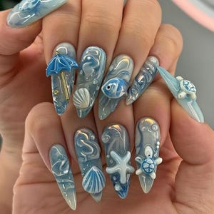 May include: A hand with long, pointed, gel nails featuring a sea theme. Each nail has a different design, including a blue umbrella, seashells, fish, a starfish, and a turtle. The nails are primarily light blue and white with some gold accents.