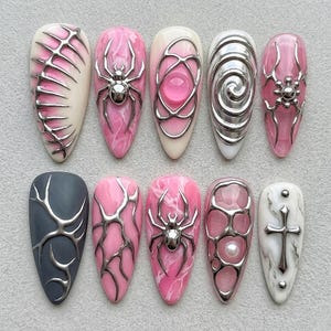 Könnte beinhalten: Eine Sammlung von zehn künstlichen Nägeln mit verschiedenen Designs. Die Nägel weisen eine Mischung aus Farben auf, darunter Rosa, Weiß und Grau, und sind mit silberfarbenen Metallverzierungen wie Spinnen, Wirbeln und geometrischen Formen verziert. Die Nägel haben ein glänzendes Finish.
