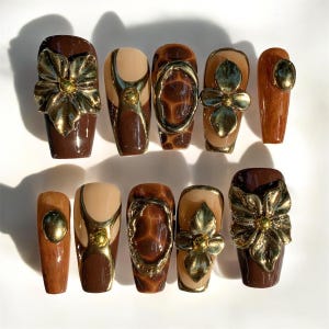 Könnte beinhalten: Ein Set aus zehn künstlichen Nägeln in verschiedenen Braun- und Beigetönen, verziert mit goldfarbenen Verzierungen. Zu den Designs gehören eine Schleife, Blumenformen und geometrische Muster. Die Nägel sind auf einer weißen Oberfläche angeordnet.