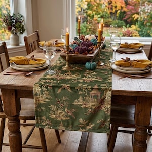 May include: A dark green velvet table runner with a gold-toned chinoiserie print featuring pagodas and birds. The runner is centered on a wooden dining table set with white plates, mustard yellow napkins, clear glassware, and brass candle holders.
