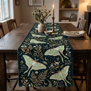 May include: A dark teal table runner featuring a repeating pattern of pale green luna moths, golden fern fronds, small white flowers, and crescent moons. The fabric has a smooth texture and covers the length of a wooden dining table.