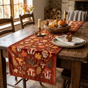 May include: A rust-colored velvet table runner featuring embroidered patterns of various mushrooms, autumn leaves, ferns, and acorns. The runner is laid on a wooden table with a wooden bowl of pumpkins and a small wooden squirrel figurine.