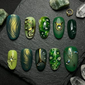 May include: A collection of ten artificial nails with various designs. The nails feature shades of green and gold, with patterns including swirls, a butterfly, and water droplet effects. Some nails have embedded gems and gold accents.