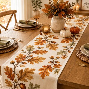 May include: A rectangular fabric table runner featuring a printed pattern of autumn oak leaves and acorns in shades of orange, brown, and green on a cream background, displayed on a wooden dining table with place settings and seasonal decor.