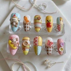 May include: A set of ten artificial nails with various designs. The nails feature cartoon monkeys, apples, fish, and patchwork patterns. Colors include light blue, pink, yellow, and brown. Each nail is displayed on a clear plastic stand.