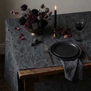 May include: A dark gray tablecloth featuring a black botanical line art pattern of ferns, leaves, and flowers. The table is set with a black plate, a metal fork, a knife, a wine glass, a black candle in a holder, and a bowl of dark flowers.
