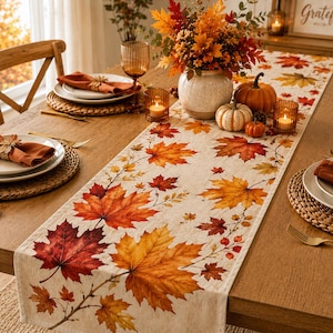 May include: A long rectangular table runner featuring a printed pattern of vibrant autumn maple leaves in shades of red, orange, and yellow on a light beige fabric. The runner is centered on a wooden table set with white plates, orange napkins, and glassware.