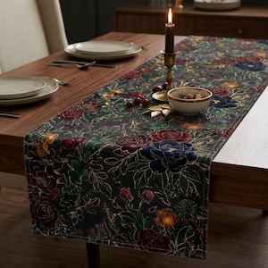 May include: A dark-colored velvet table runner featuring a floral pattern with red, blue, and yellow flowers, green leaves, and gold-colored outlines. The runner is placed on a wooden table with white plates, a candle in a holder, and a small bowl.