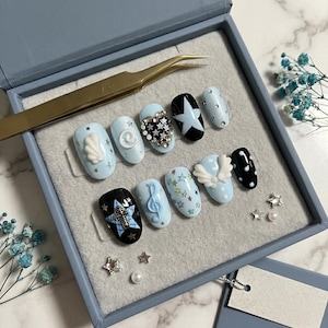 May include: A set of light blue press-on nails with various designs, including stars, a seashell, and musical notes. Some nails feature rhinestones and a black and white star design. The set is displayed in a blue box with a pair of tweezers.
